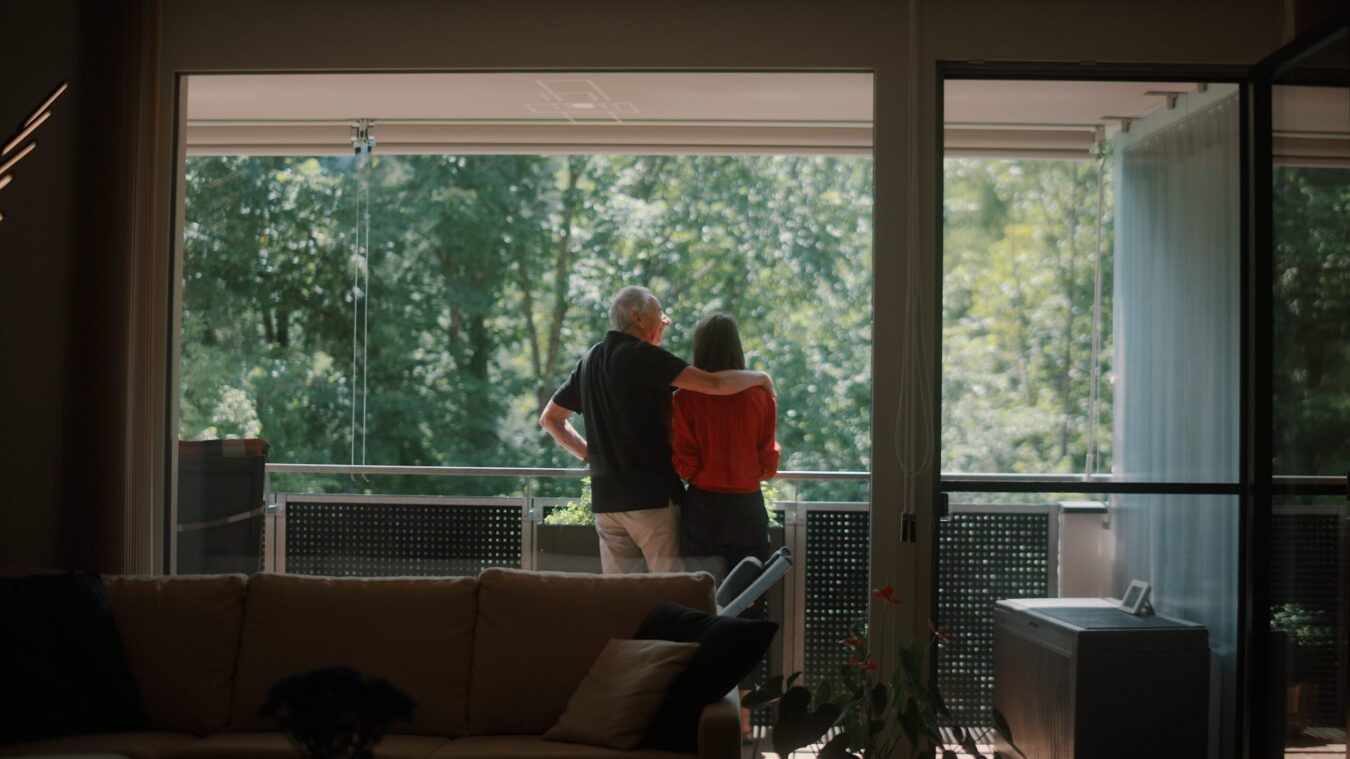 Ehepaar auf dem Balkon ihrer Wohnung, Mann legt Arm um seine Frau, sie blicken ins Grüne