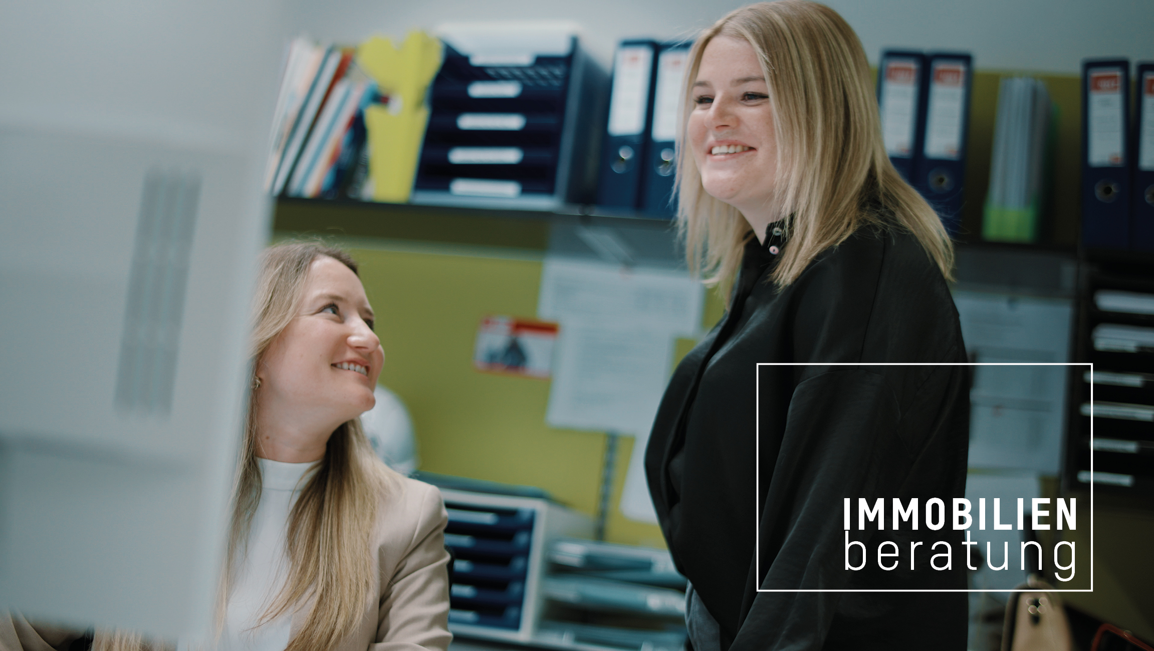 Zwei blonde Frauen in einem Büro, eine sitzt am Computer und schaut die andere an, die andere steht daneben und schaut in Computer