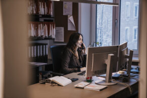 Eine Frau mit braunen langen Haaren im Büro an ihrem Schreibtisch, telefoniert, im hintergrund ein Ordnerregal