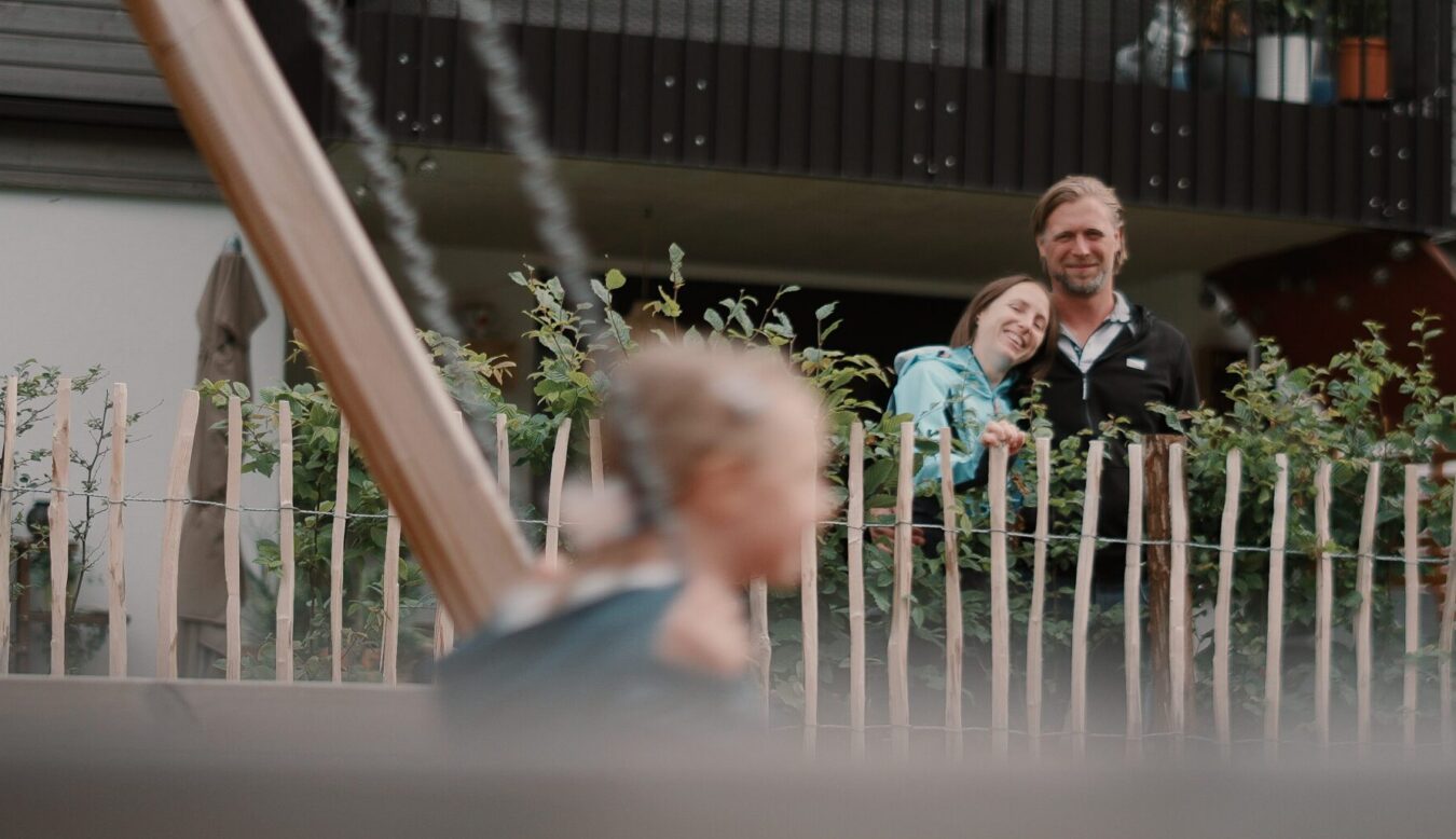 Garten einer Wohnung, Kind schaukelt im Vordergrund, ist aber verschwommen, Eltern schauen vom Gartenzaun zum Kind