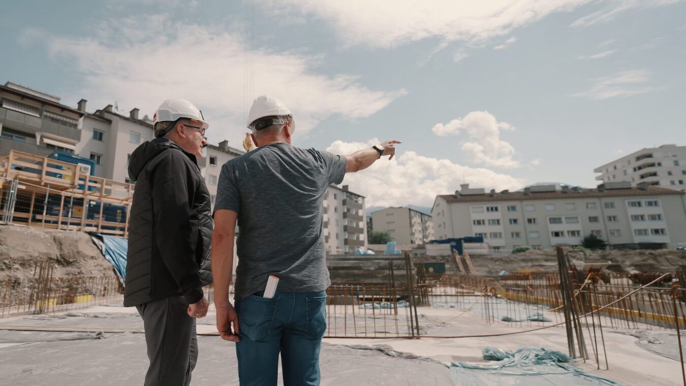 Zwei Männer auf Baustelle, mit Bauhelmen, einer zeigt auf Baustelle