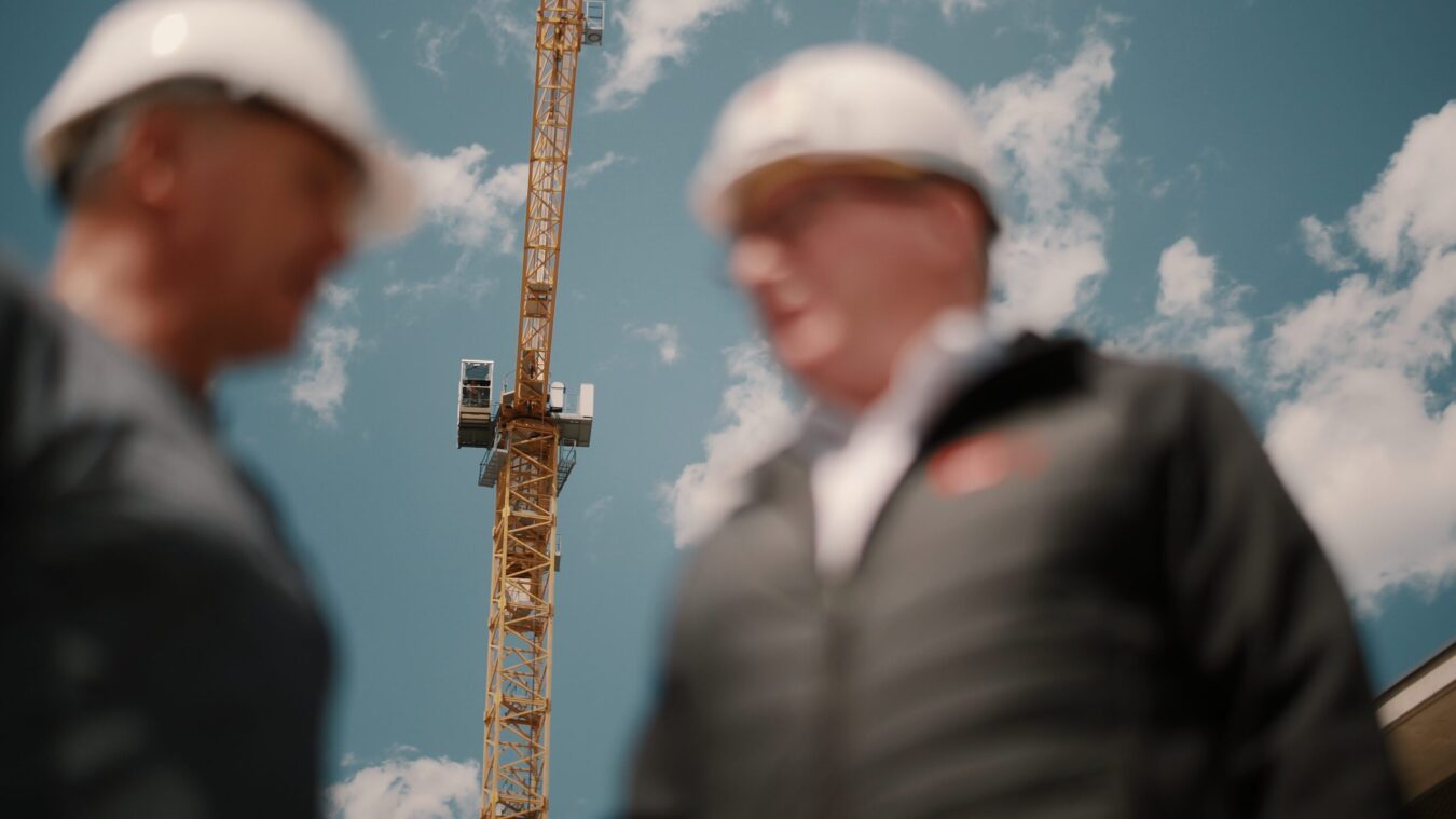 Zwei Männer vor einem Baukran, Männer nur mit Oberkörper und Gesicht, mit Bauhelm, reden miteinander, sind aber unkenntlich verschwommen, Kran im hintergrund ist scharf zu sehen