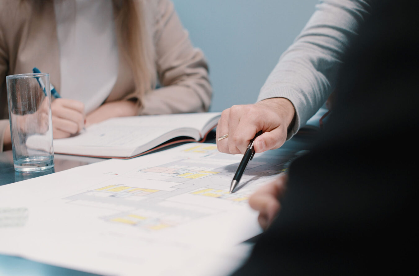 Meetingsituation, man sieht keine GEsichter, eine Hand zeigt mit Stift auf ein Schriftstück