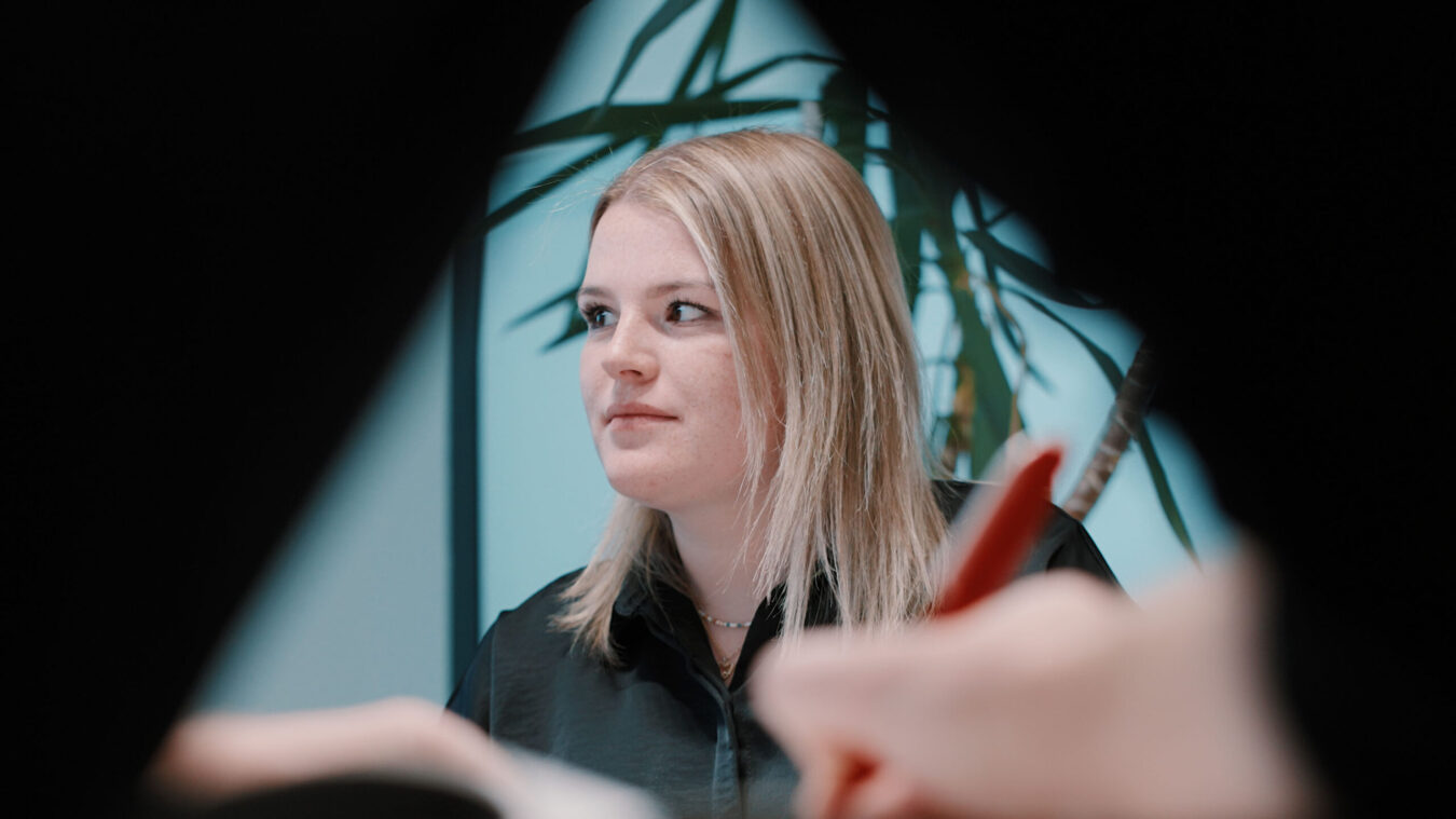 Eine blonde junge Dame schaut auf einen Gesprächspartner, der nicht im Raum ist, im Vordergrund eine Hand mit rotem Stift