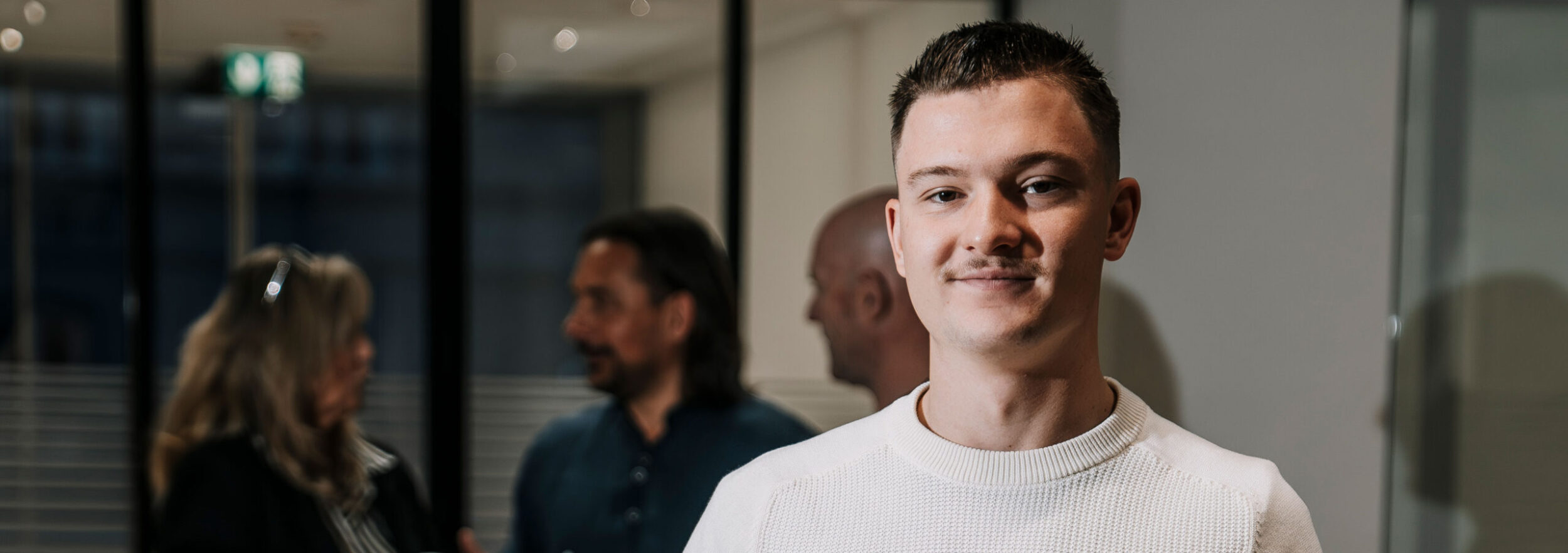 ein junger Mann im Vordergrund, im Hintergrund verschwommen weitere Personen ,die sich unterhalten